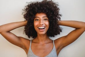 Black woman with healthy hair and putting her hands on her head while smiling with a big, bright and beautiful smile.